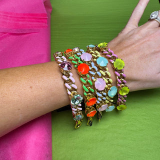 A close-up of a person's wrist wearing a stack of chunky gold-tone chain bracelets embellished with colorful faux gemstones in pink, orange, green, blue, and purple, set against a split pink and green background. TOVA