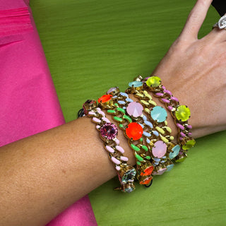 A close-up of a person's wrist wearing a stack of chunky, gold-tone bracelets heavily decorated with colorful faux gemstones in pink, orange, green, blue, and purple, set against a bright green and pink background. TOVA