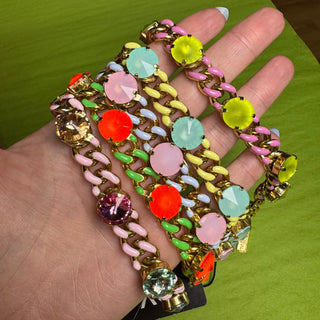 A close-up of a hand holding a stack of colorful chain bracelets decorated with large faceted gemstones in pink, orange, yellow, green, and blue, set against a bright green background. TOVA