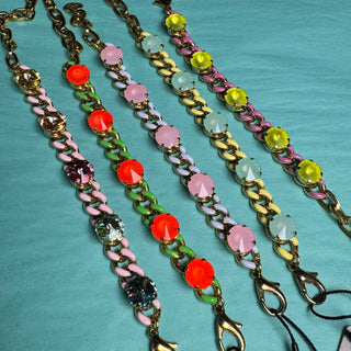 Four Tova brand beaded chain bracelets with gold-tone hardware and colorful faceted beads, laid out on a teal fabric background with their retail tags visible. TOVA