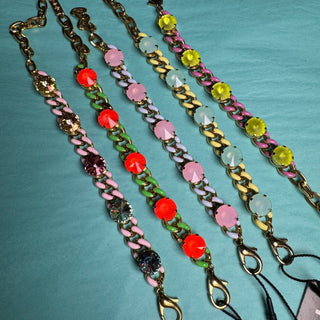 Four colorful chain-link wristbands with faceted gemstone accents are laid out on a teal background. The wristbands feature gold-tone chains and links, with pink, red, green, yellow, and clear/light blue stones. TOVA