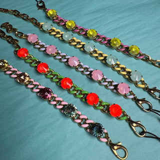 Four colorful chain link bracelets with faceted rhinestones in pink, red, yellow, green, and blue, laid out on a light blue textured surface. A black tag with 'TOVA' is visible at the top. TOVA