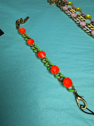 A gold-tone chain bracelet featuring alternating bright orange-red oval beads and light green oval links, laid out on a turquoise fabric background.