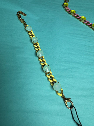 A gold-tone chain bracelet with round, pale blue-green cabochon stones, laid diagonally on a bright turquoise fabric background.