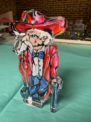 A hand-painted acrylic figurine of colonel reb in a red suit and hat with a blue bow tie and blue pants, standing on a green surface with a brick wall in the background.