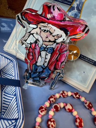 A hand-painted acrylic figurine of a cowboy in a red hat and jacket, displayed with a blue and white patterned bag, a beaded necklace, and printed cards on a blue surface.