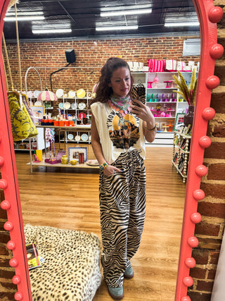 A woman takes a mirror selfie in a boutique, wearing a cream sleeveless vest, a colorful graphic t-shirt, and zebra-print wide-leg pants. The boutique has a brick wall, wooden floors, and a large pink-framed mirror.