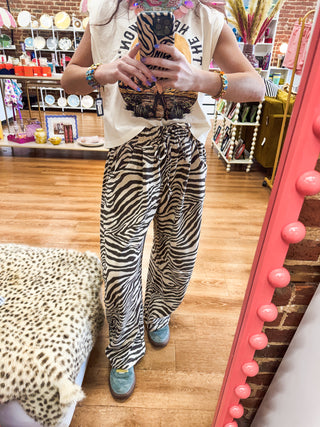 A person standing in a boutique mirror wearing a cream graphic sleeveless top and wide-leg zebra-print pants, with blue shoes, in a shop with wood floors and shelves in the background.