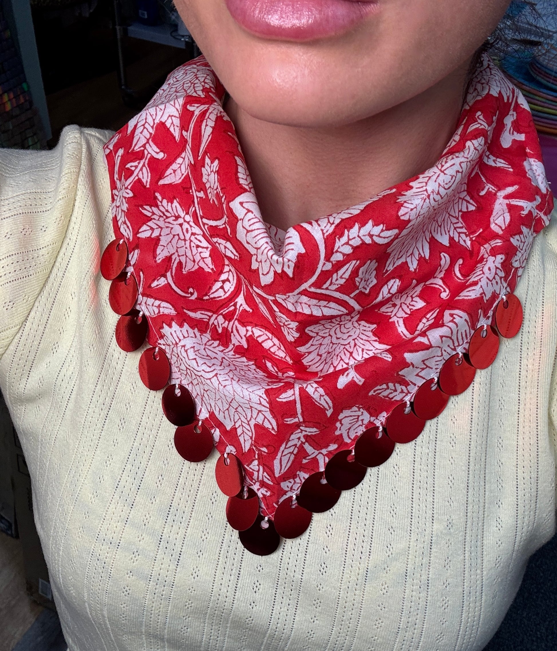 A close-up of a person wearing a red and white floral patterned neck scarf with a row of red circular sequins along the bottom edge, paired with a cream ribbed knit top.