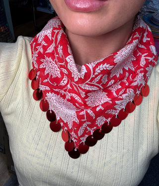 A close-up of a person wearing a red and white floral patterned neck scarf with a row of red circular sequins along the bottom edge, paired with a cream ribbed knit top.