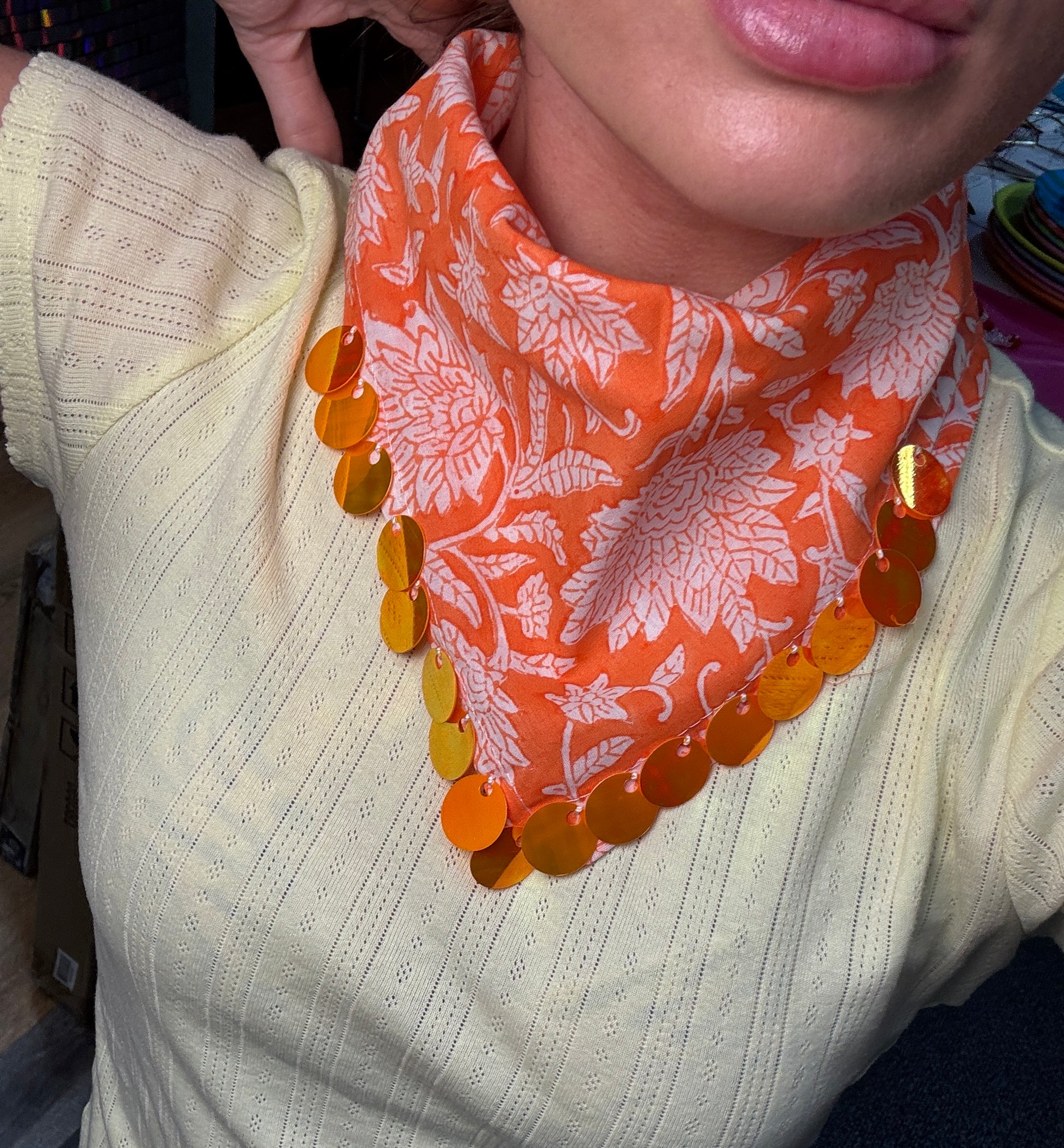 An orange and white floral patterned neck gaiter or bandana with shiny metallic gold sequins along the edges, worn by a person in a pale yellow knit sweater.
