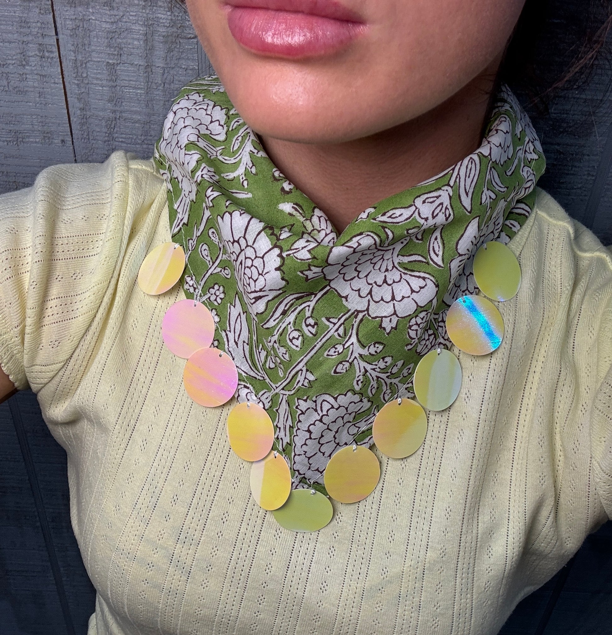 A close-up of a person wearing a green and white floral patterned neck scarf with a row of iridescent oval beads draped over a pale yellow ribbed top.