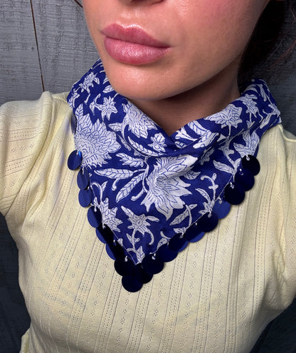 A close-up of a person wearing a navy blue and white floral bandana-style neck scarf with dark blue pom-pom trim, paired with a cream ribbed top.