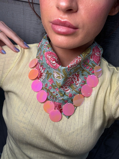 A close-up of a person wearing a chunky statement necklace with iridescent pink and peach circular discs layered over a floral neck scarf and a pale yellow ribbed sweater.