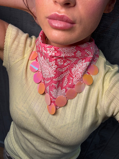 A close-up of a person wearing a pink paisley bandana-style neck scarf with a row of round, disc-like embellishments in pink, peach, and orange, paired with a pale yellow ribbed top.
