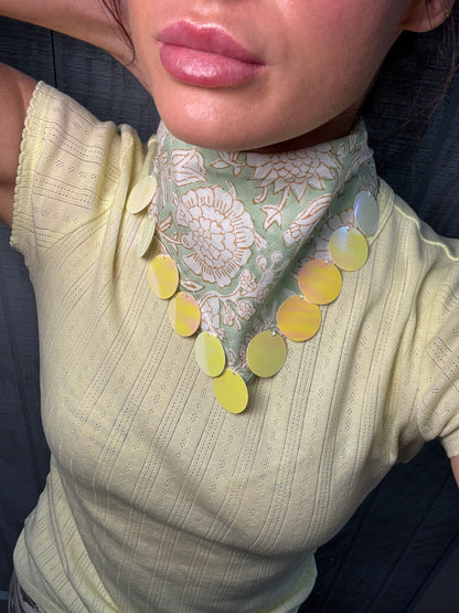 A close-up of a person wearing a pale yellow ribbed knit top and a patterned neck scarf with large iridescent yellow-green oval discs around the neck.