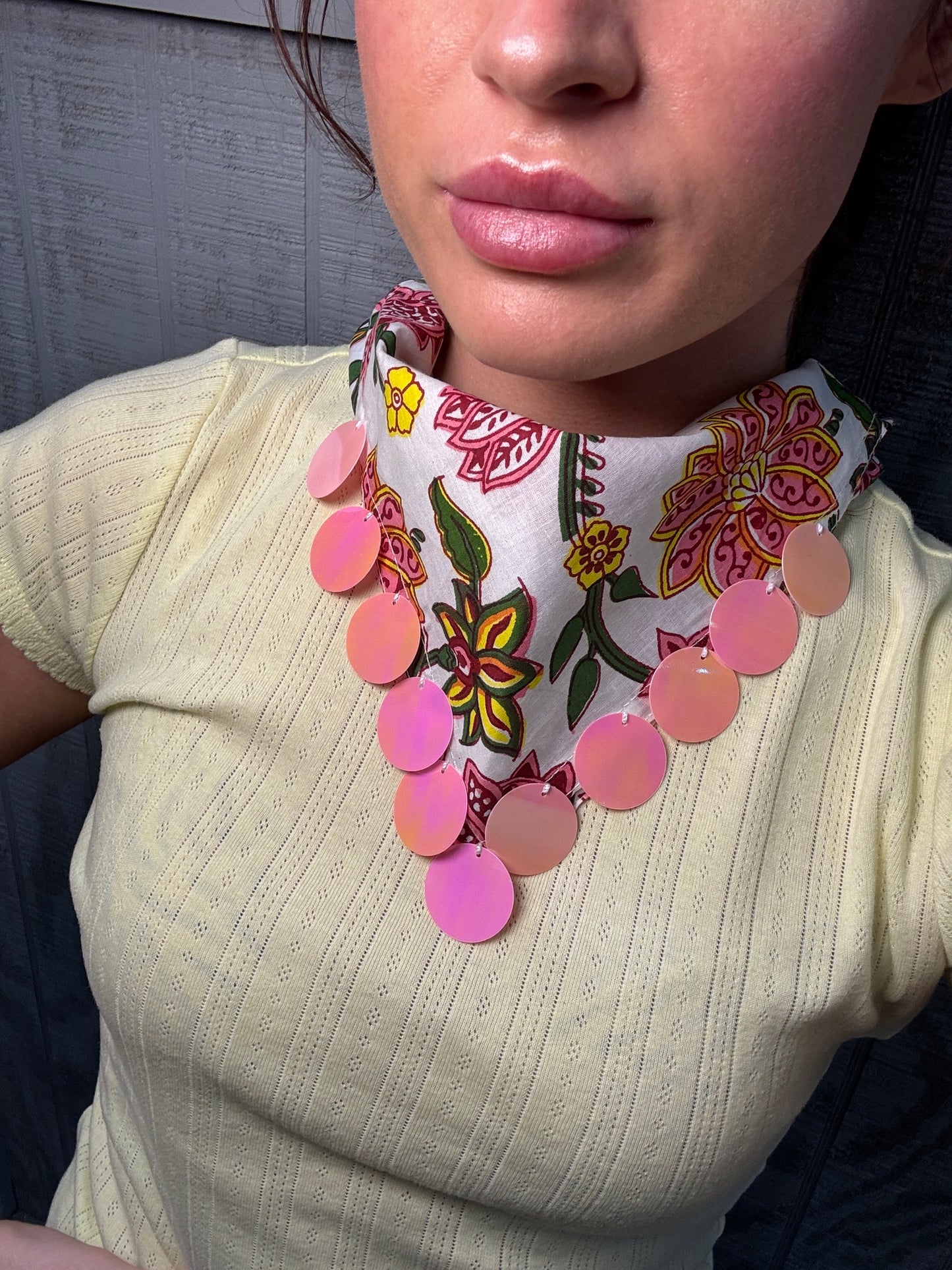 A close-up of a person wearing a chunky statement necklace with a white floral collar and pink circular disc accents, paired with a pale yellow ribbed top.