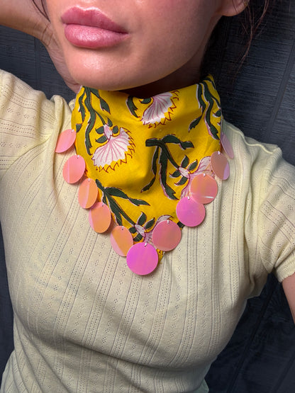 A close-up of a person wearing a bright yellow neck scarf with a black and white floral pattern and a row of pink circular disc charms, paired with a pale yellow ribbed top.
