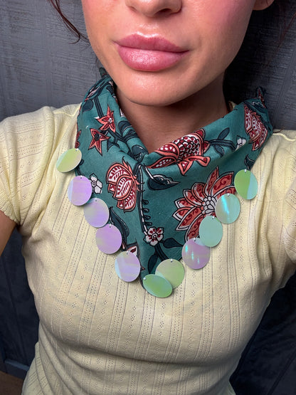 A close-up of a person wearing a teal patterned neck scarf with red and white floral motifs and a row of iridescent oval beads in pastel green, lavender, and mint over a cream ribbed top.