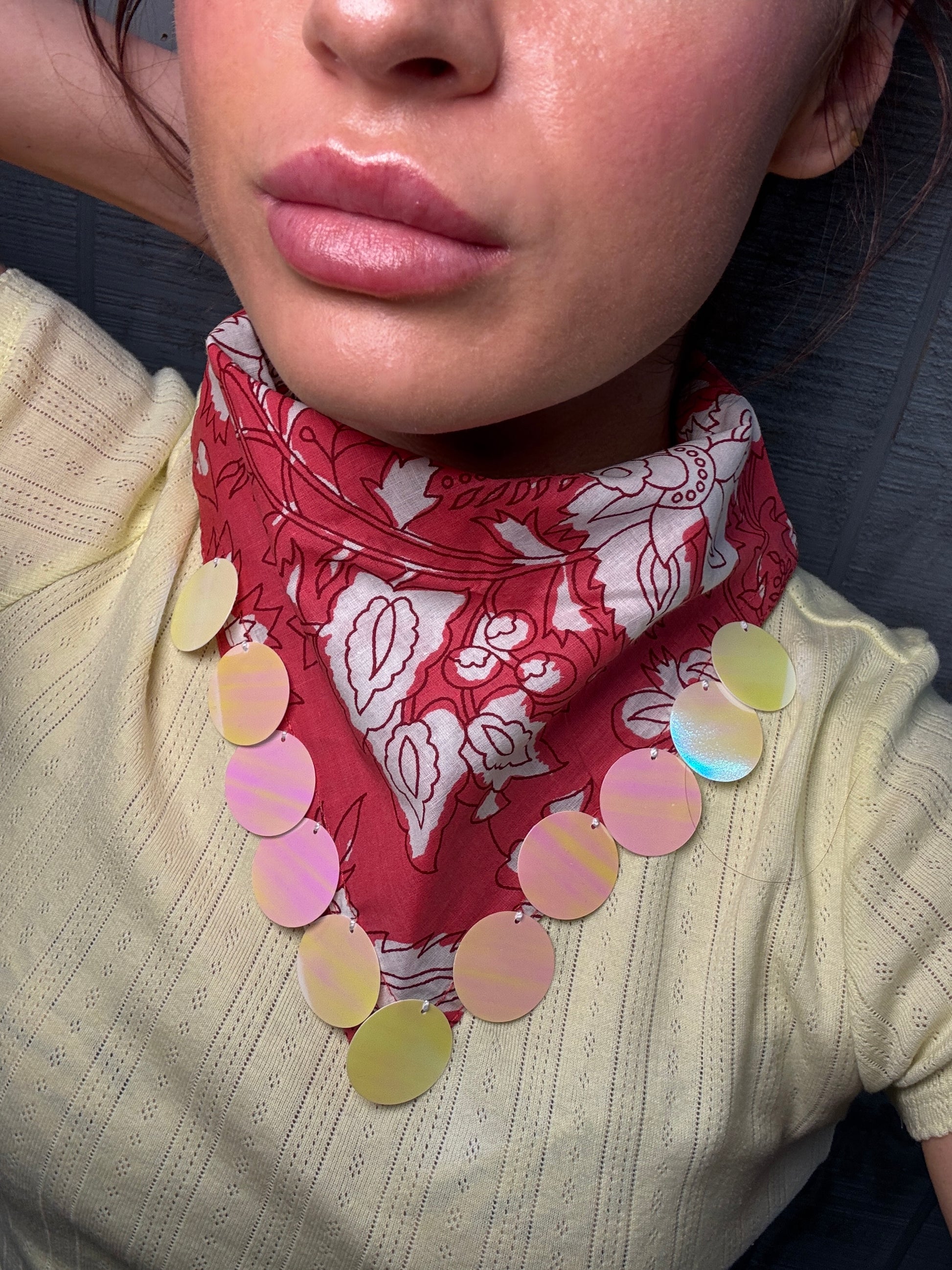 A close-up of a person wearing a red and white patterned neck scarf with a necklace of iridescent circular discs draped over the shoulders, paired with a pale yellow ribbed knit top.
