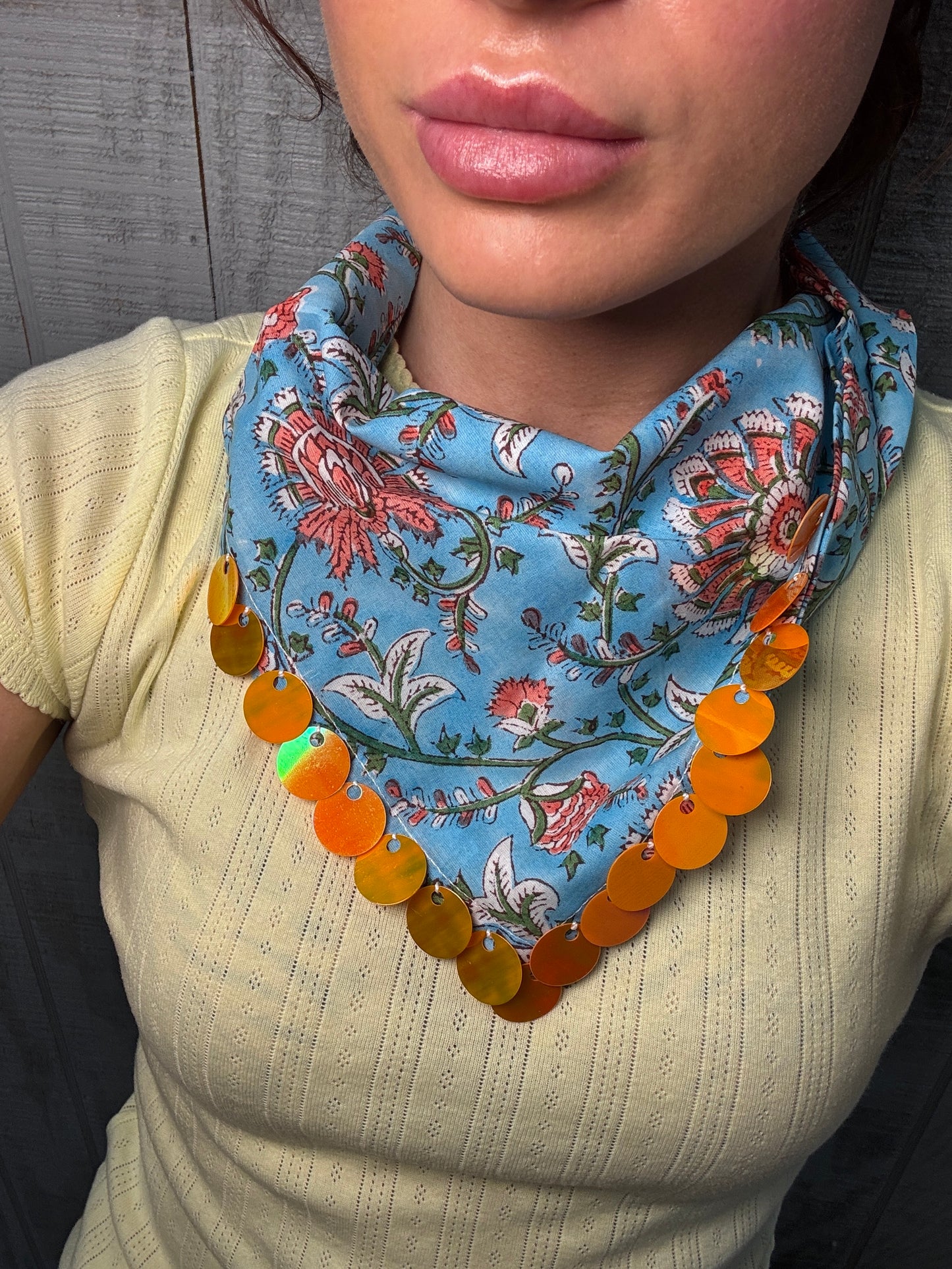 A close-up of a person wearing a light blue floral neck scarf with a row of shiny orange circular disc charms, paired with a cream ribbed knit top against a dark wood background.