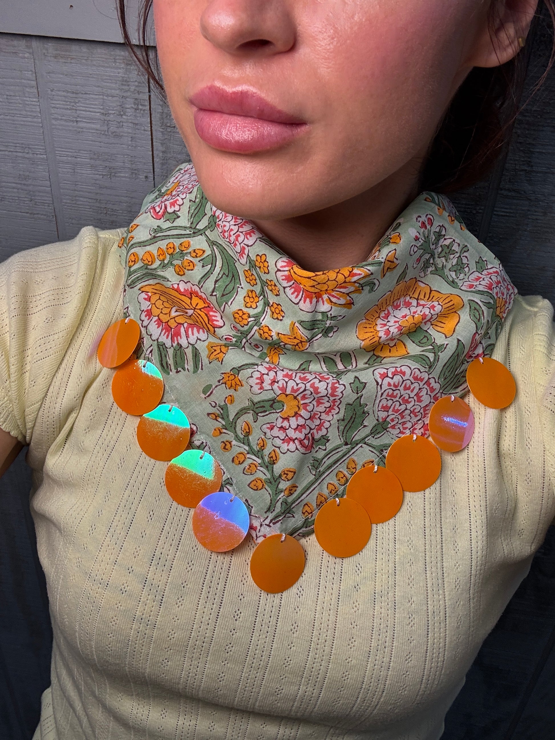 A close-up of a person wearing a cream ribbed knit top and a patterned bandana-style neck scarf with orange circular disc accents.