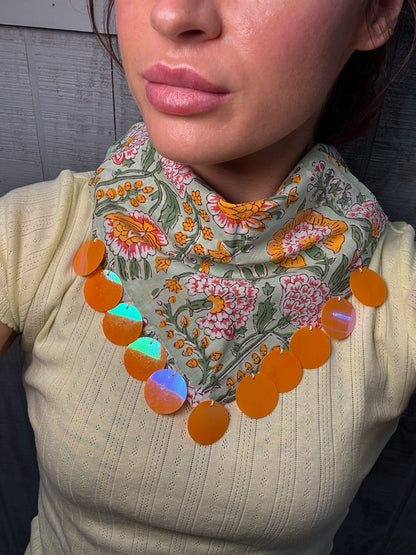 A close-up of a person wearing a cream ribbed knit top and a patterned bandana-style neck scarf with orange circular disc accents.