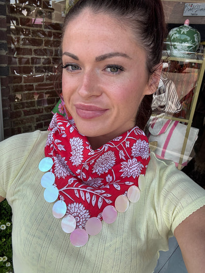 A woman wearing a red and white floral print neck scarf with large iridescent shell-like circular accents, paired with a pale yellow ribbed top. She is taking a selfie in front of a shop window with a brick wall visible.