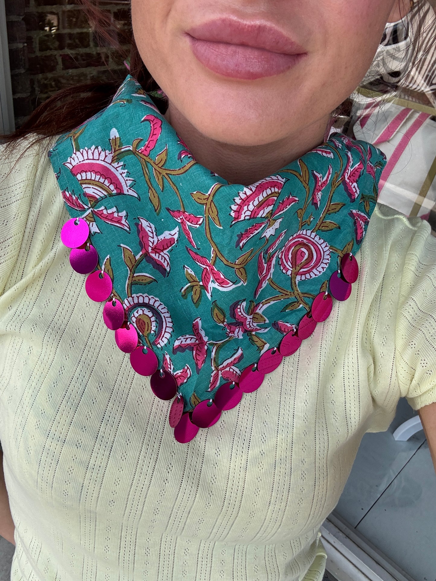 A close-up of a person wearing a teal bandana-style necklace with a pink, white, and gold floral print and a row of shiny magenta sequins along the edge, paired with a pale yellow ribbed top.