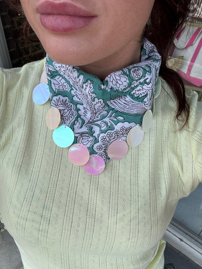 A close-up of a person wearing a green and white patterned neck scarf with a bib-style collar, adorned with multiple round pastel-colored shell-like disc charms, paired with a pale yellow ribbed top.
