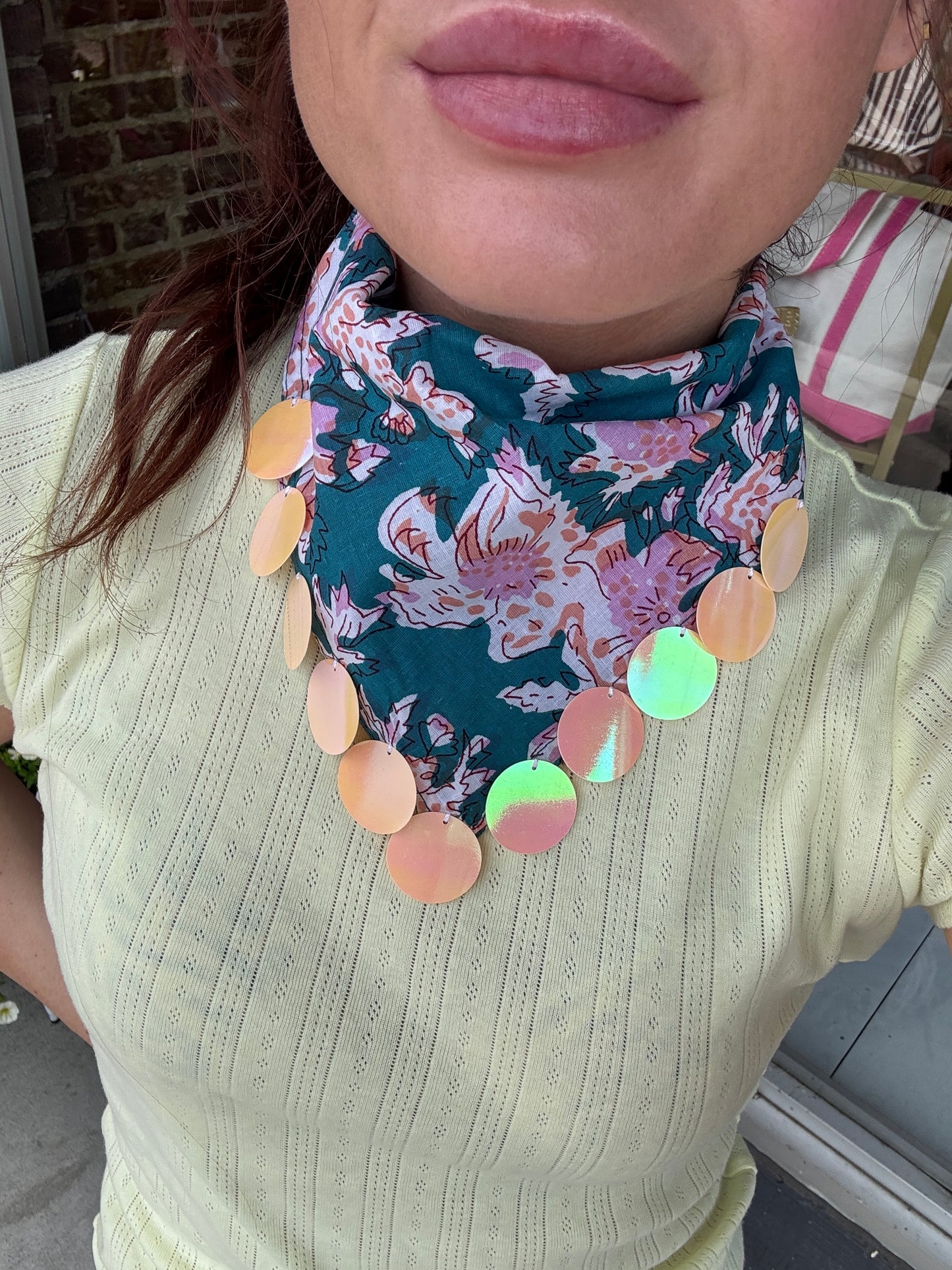 A close-up of a person wearing a teal bandana-style neck scarf with a pink floral print and a row of iridescent peach-colored circular discs around the edge.