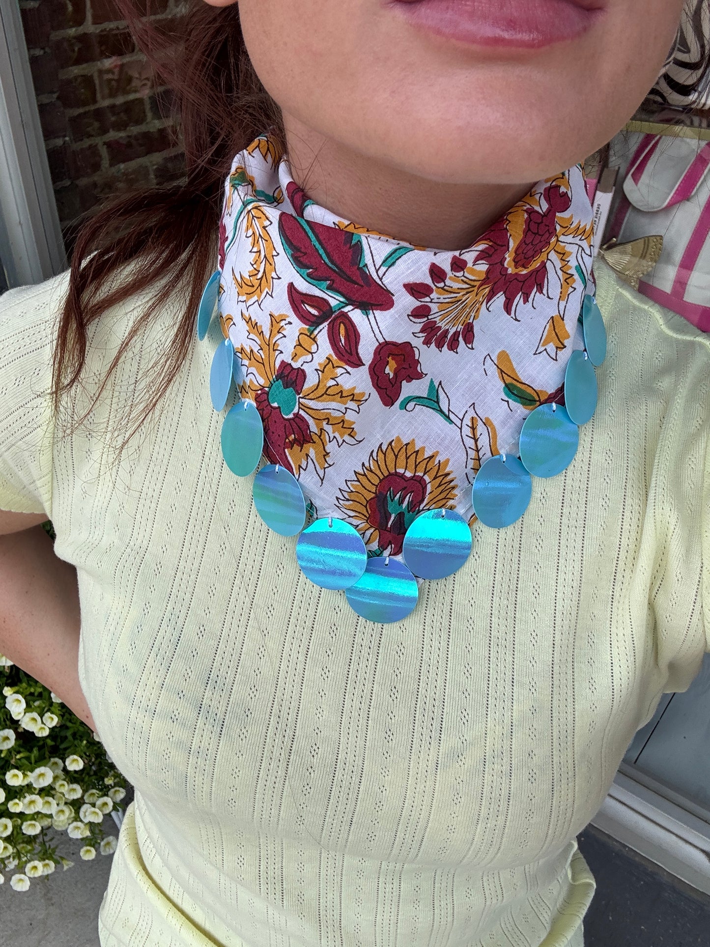 A close-up of a person wearing a white neck scarf with a colorful floral and sunflower print, accented by large iridescent blue-green circular beads at the bottom.
