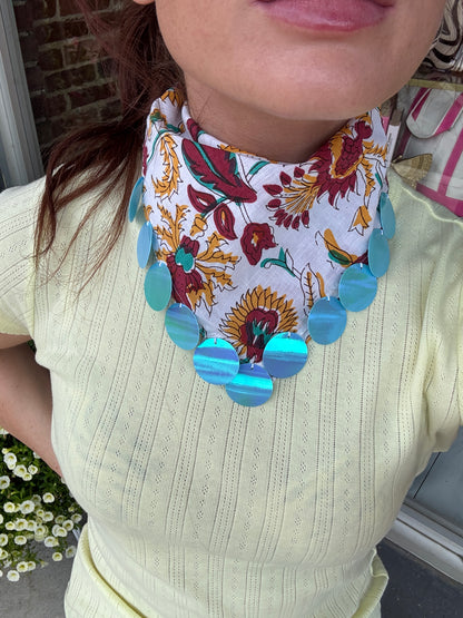 A close-up of a person wearing a white neck scarf with a colorful floral and sunflower print, accented by large iridescent blue-green circular beads at the bottom.