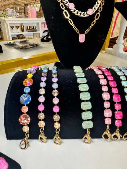 A display of colorful beaded bracelets with gold-tone clasp ends, arranged on black velvet jewelry stands alongside a gold chain necklace with pink beads on a mannequin bust. The bracelets feature various bead patterns including round, rectangular, and striped designs in pink, blue, green, purple, and orange. TOVA
