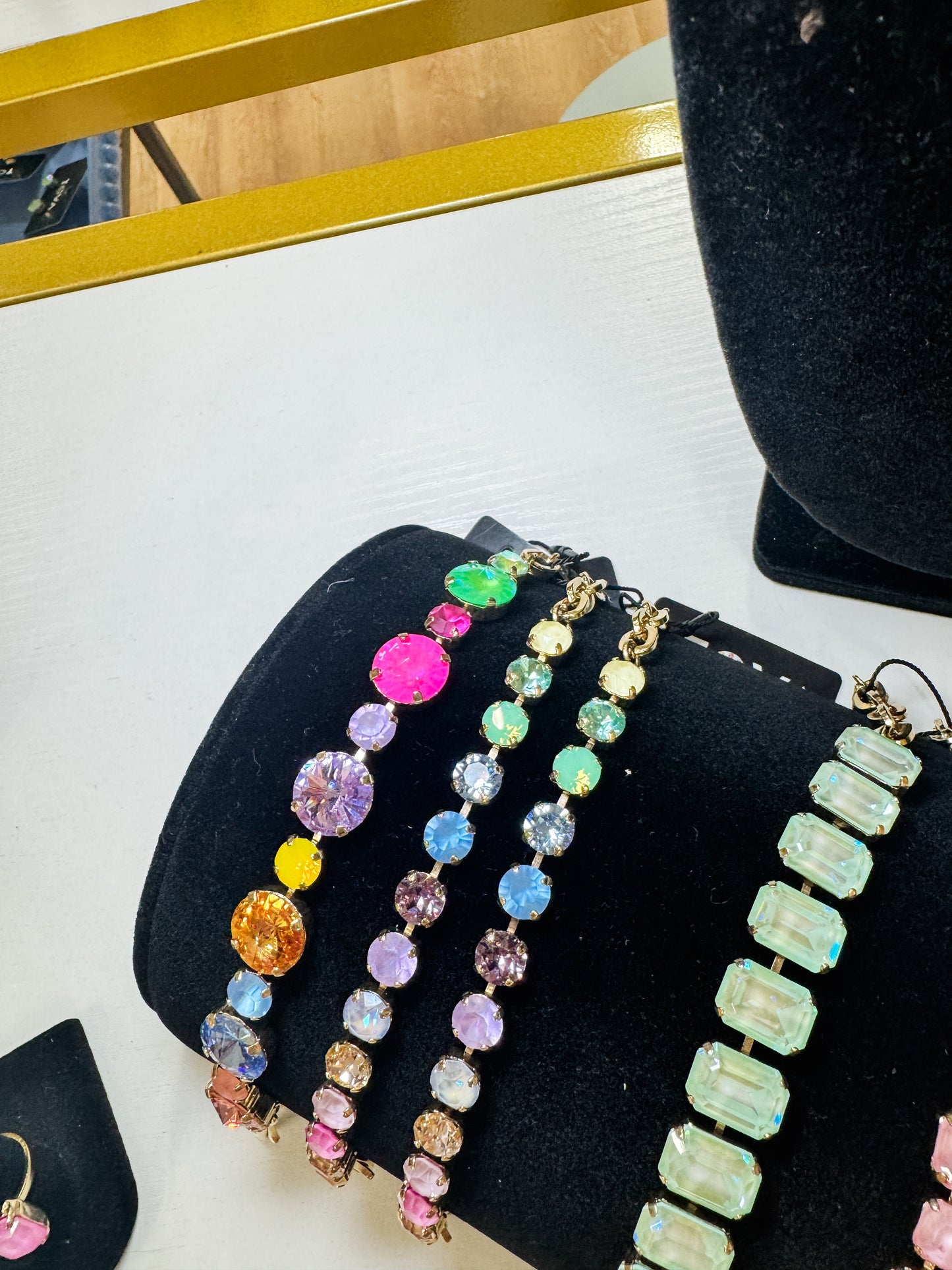 A close-up of several colorful beaded bracelets displayed on a black jewelry bust. The bracelets feature round and rectangular faceted rhinestones in vibrant colors including pink, purple, blue, green, orange, and gold-tone metal settings, arranged on a white tabletop with a gold-framed mirror in the background.TOVA