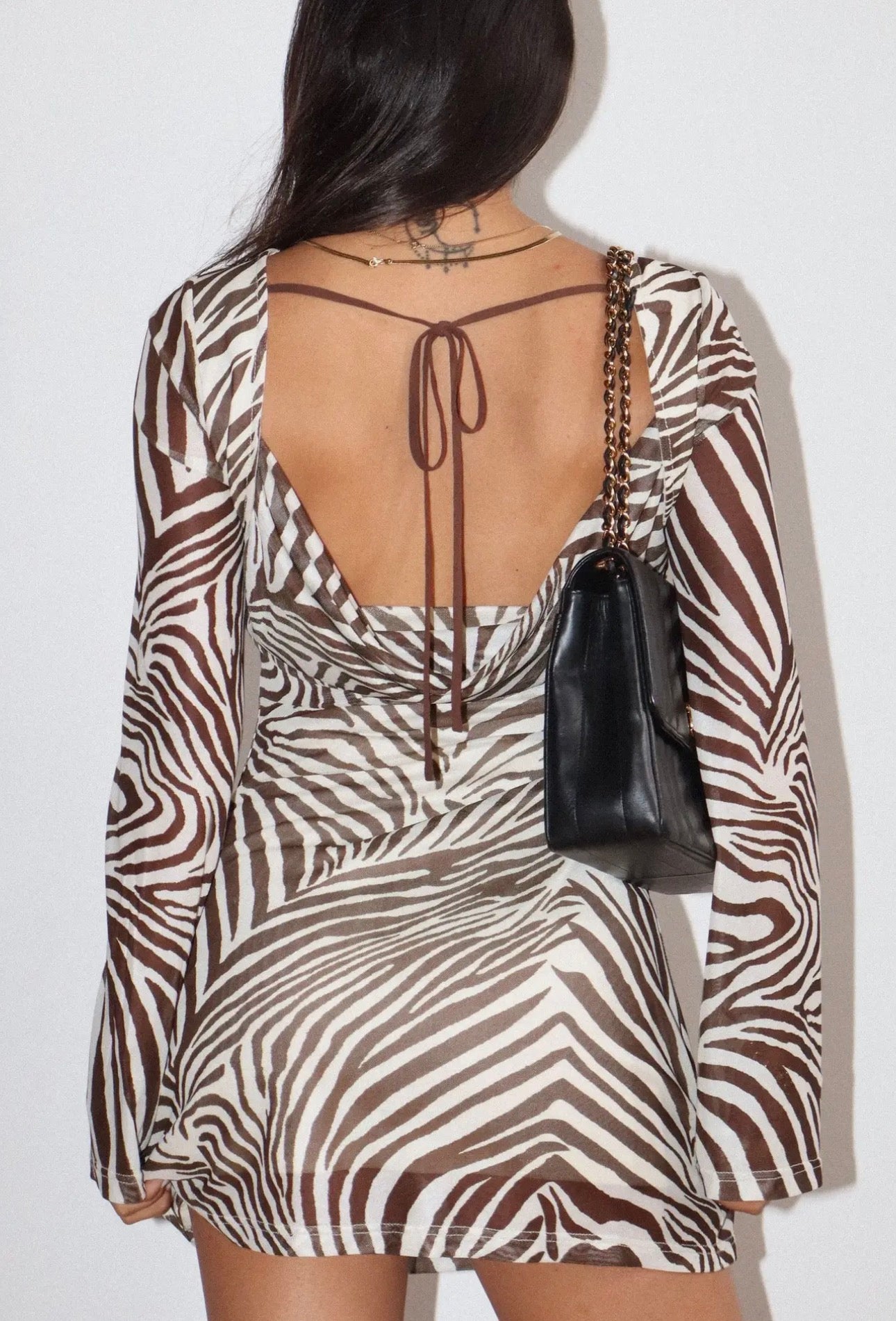 A woman shown from the back wearing a long-sleeve zebra-print dress with a deep open back and tie detail, carrying a small black handbag against a plain light background.