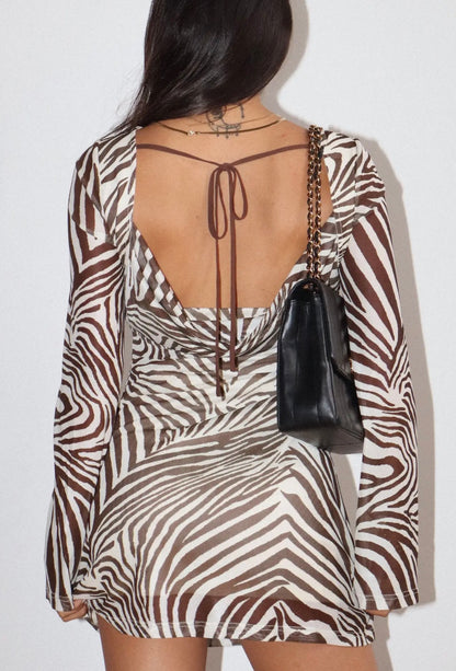 A woman shown from the back wearing a long-sleeve zebra-print dress with a deep open back and tie detail, carrying a small black handbag against a plain light background.