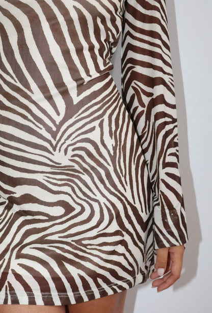 A close-up of a long-sleeve mini dress featuring a brown and off-white wavy zebra stripe pattern, worn by a model whose hand is visible on the right.