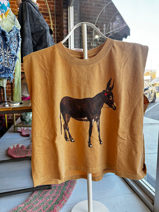 A mustard yellow t-shirt on a white hanger stand, featuring a graphic print of a dark brown donkey. The shirt is photographed in a shop window setting with other clothing and a brick wall visible in the background.