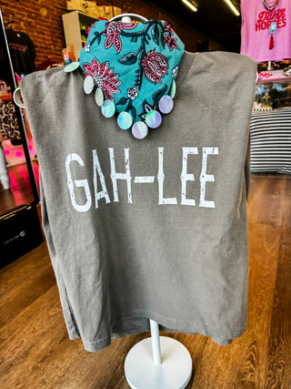 A gray graphic T-shirt displayed on a white stand in a retail store, featuring the text 'GAH-LEE' in distressed white letters. A colorful floral bandana with hanging iridescent pom-poms is draped over the collar area.