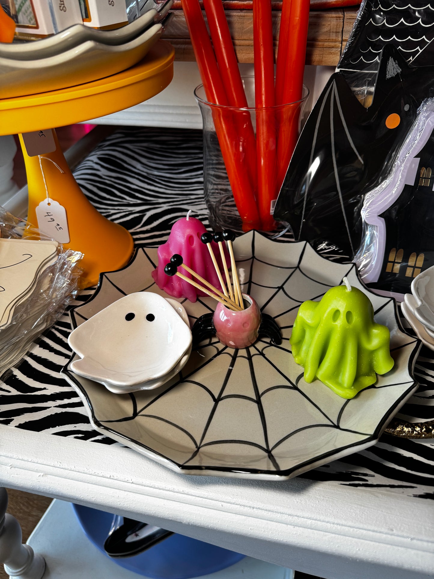 A Halloween-themed party spread on a shelf, featuring a spiderweb platter with a white ghost dish, a pink ghost candle, a green ghost candle, and a small black spider, alongside red straws and a black bat-shaped decoration.