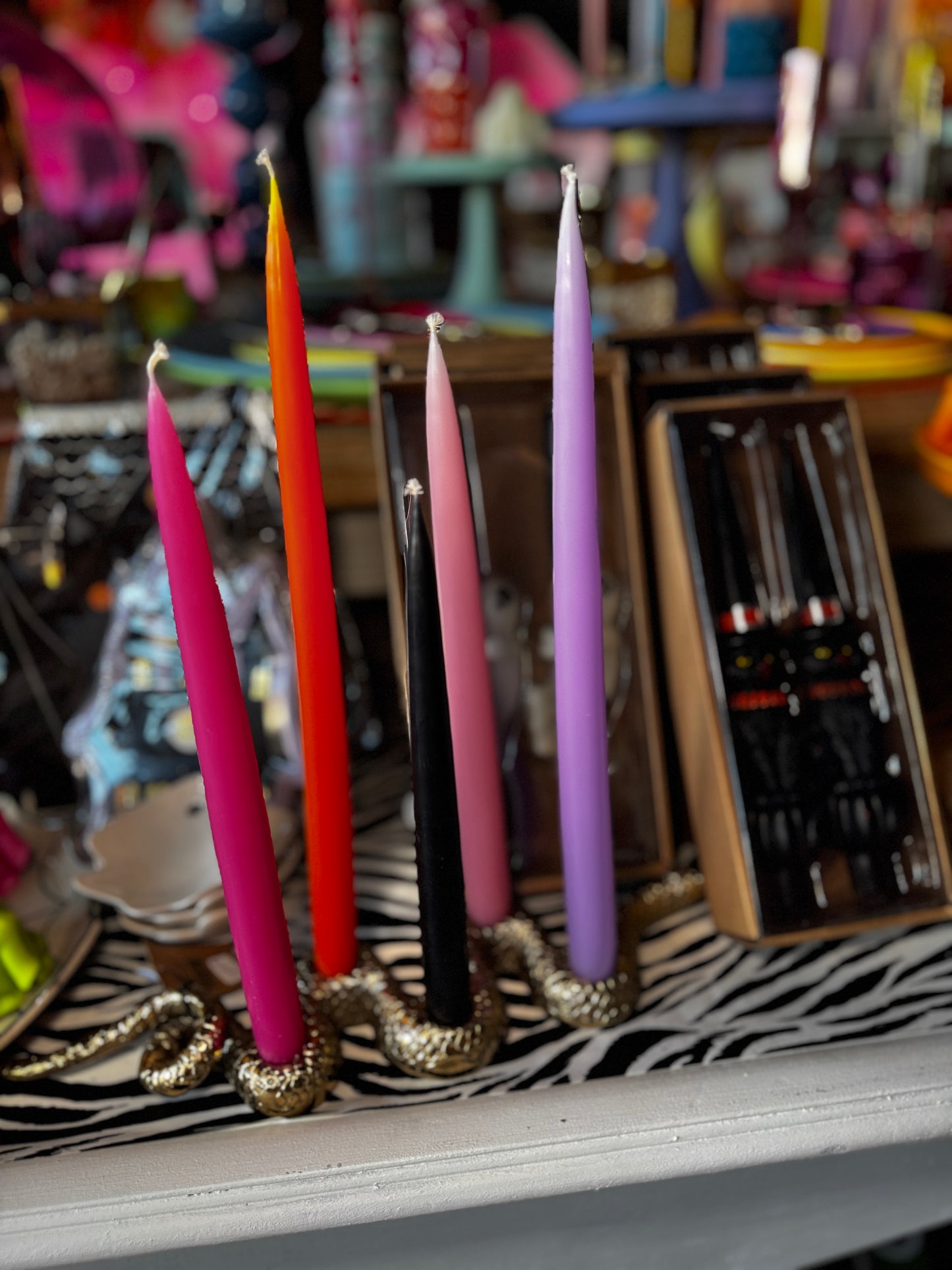 Five taper candles in pink, orange, black, light pink, and purple, displayed on a metallic gold snake-shaped holder over a zebra-print surface in a colorful shop setting.