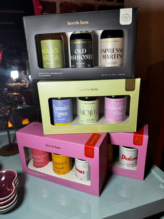 Three boxed sets of Here's How cocktail mixers stacked on a tabletop. The top set is in a black box with three dark bottles and a yellow-green label.