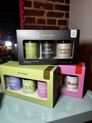 Three boxed canneries from Here's How are stacked on a reflective white surface against a brick wall background. The top black box contains three dark glass cans labeled Margari, Old Fashioned, and Espresso Martin.