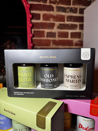 A boxed cocktail mixer set from Here's How, containing three bottles: Margarita, Old Fashioned, and Espresso Martini, presented in a black gift box. The box is stacked on top of other colorful product boxes against a brick wall background.