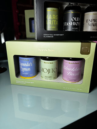 Two boxed cocktail mixer sets from here's how are stacked on a reflective white surface. The front set is in a light green box with three bottles visible: lavender lemon, mojito, and blackberry basil.