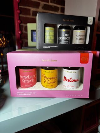 Two boxed beverage gift sets from herb's how stacked on a light surface. The front set is a pink box containing three small bottles labeled Strawberry Smash, Pineapple Ranch, and Paloma.