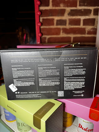 A black rectangular product box with printed text on the back, stacked on top of other colorful boxes including a lime green one and a pink one. The main box is shown from the back, with a brick wall and a red-framed mirror visible in the background.