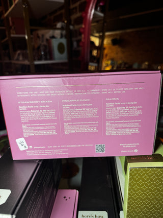 The back of a pink cosmetic box on a store shelf, showing product names and text in white print. Visible text includes Strawberry Smash, Pineapple Punch, and Paloma.