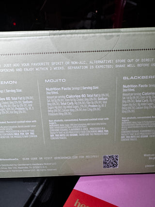 Close-up of the back of a beverage box showing flavor labels for Lemon, Mojito, and Blackberry, with nutrition facts and a QR code. A pink item is partially visible underneath.
